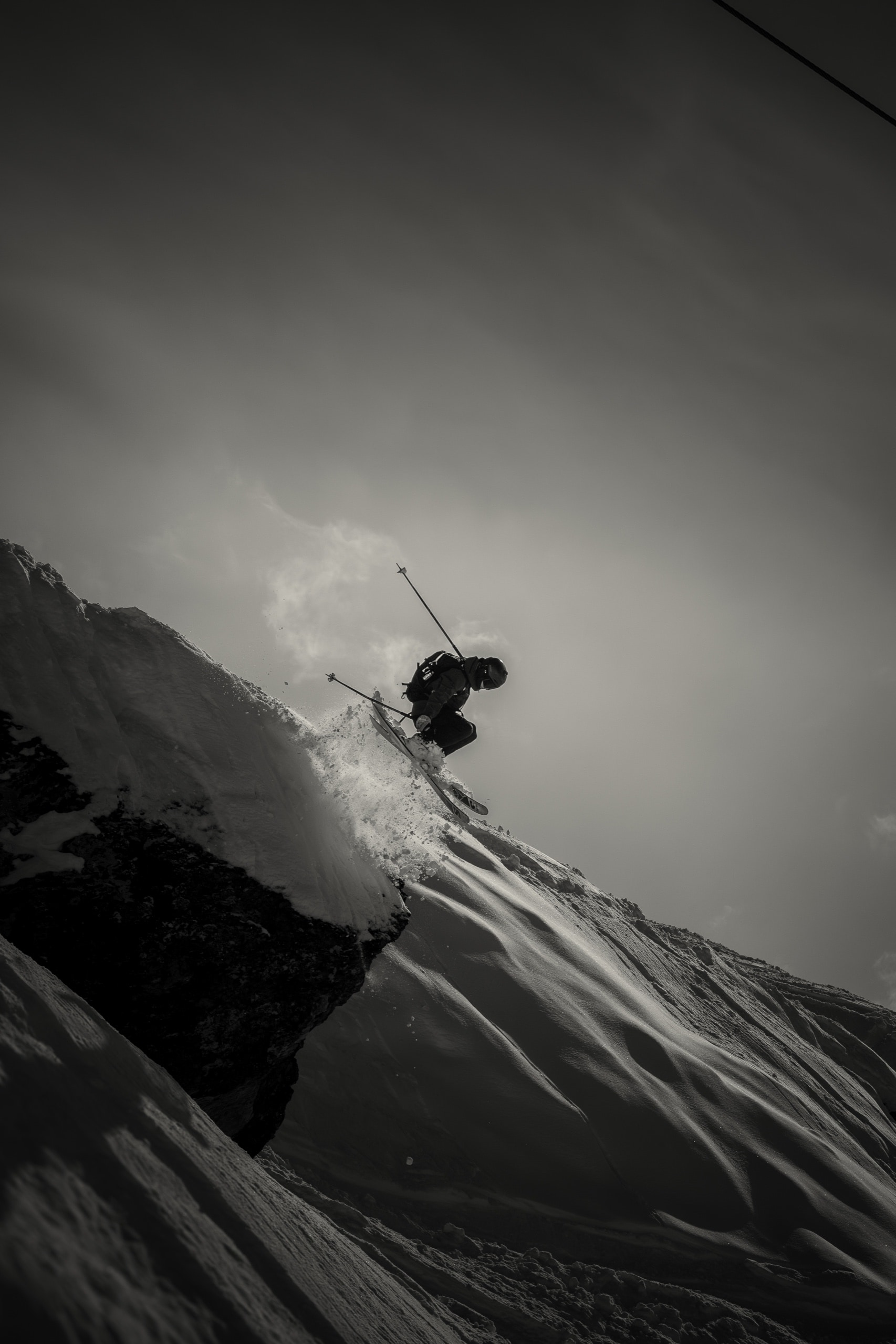 Silhouette of a skier jumping off a snowy cliff against a bright mountain sky, in black and white