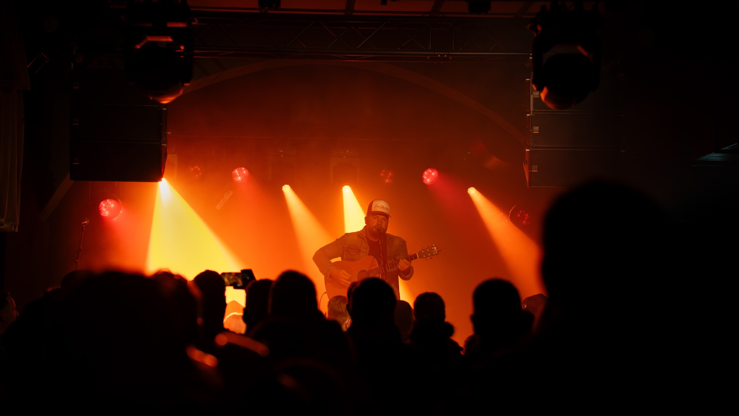 Musician playing acoustic guitar on an orange-lit stage, viewed from behind a concert crowd