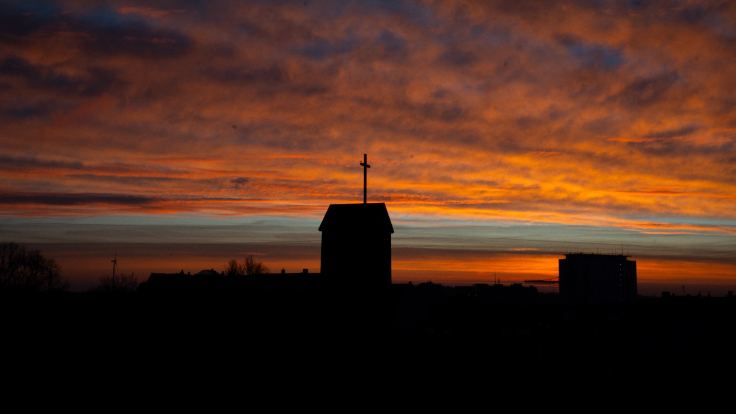 Silhouette of a church steeple with a cross against a fiery sunset sky over a town skyline