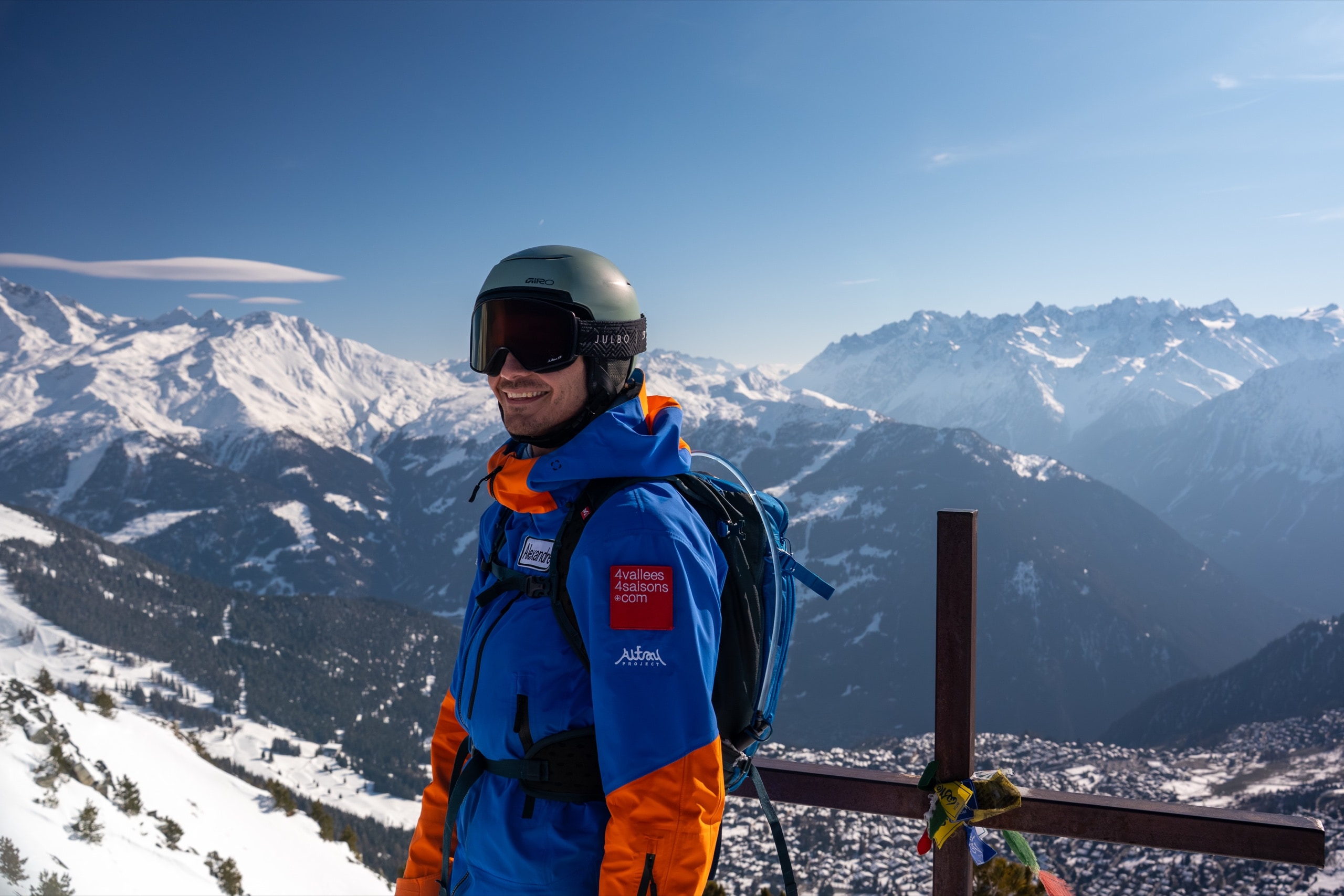 Skier in a blue and orange jacket smiling on a snowy mountain summit with alpine peaks behind