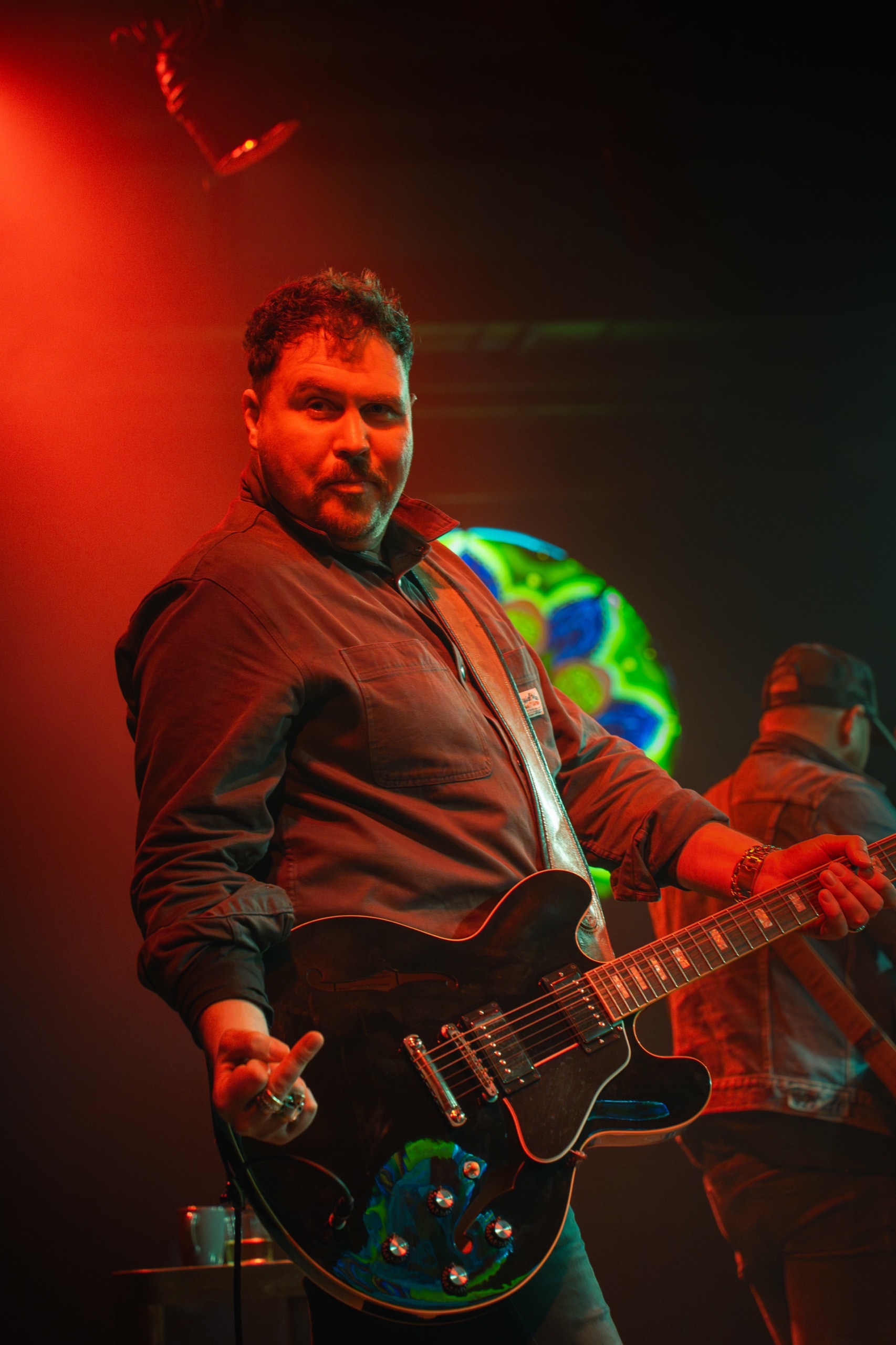 Two musicians on stage—one smiling in a denim jacket and trucker hat playing guitar, another silhouetted in the foreground singing