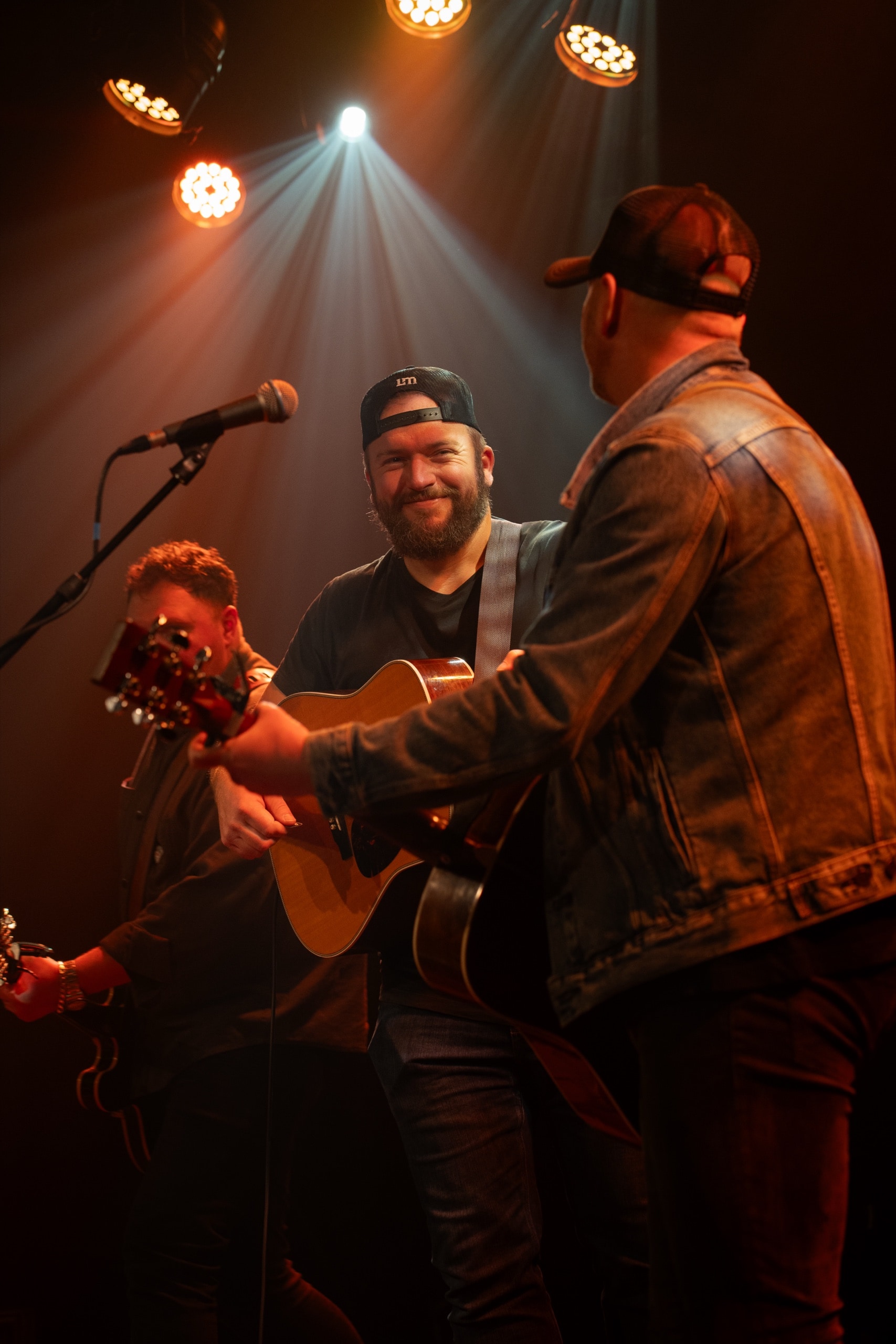 Bearded musician smiling while playing acoustic guitar on stage under warm spotlights with bandmates